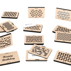 May include: A collection of nine wooden gift card holders, each featuring a unique laser-cut design. The designs include geometric patterns, hearts, and floral motifs. The holders are crafted from natural wood and have a rustic, handmade appearance. Two of the holders display the text "Happy Birthday" and "Gift Card".