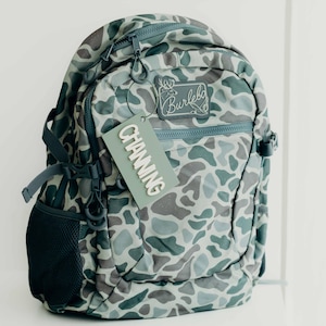 Puede incluir: Una mochila con estampado de camuflaje en tonos grises, verdes y azules. La mochila tiene m&uacute;ltiples compartimentos, cremalleras y un bolsillo lateral de malla. Una etiqueta que dice "CHANNING" cuelga de la cremallera. Tambi&eacute;n es visible la marca "Burlebo".