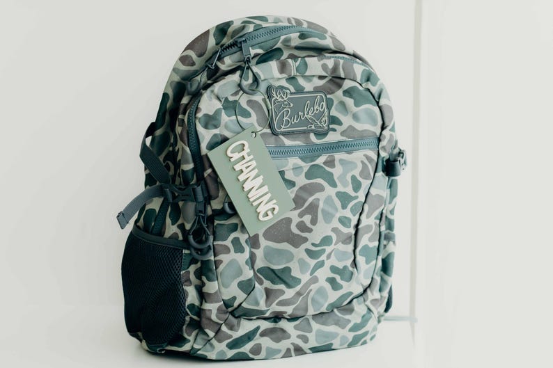 Puede incluir: Una mochila con estampado de camuflaje en tonos grises, verdes y azules. La mochila tiene m&uacute;ltiples compartimentos, cremalleras y un bolsillo lateral de malla. Una etiqueta que dice "CHANNING" cuelga de la cremallera. Tambi&eacute;n es visible la marca "Burlebo".