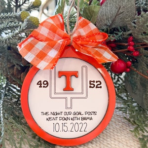 May include: Orange and white checkered bow on a round orange ornament with a white background. The ornament has a black outline of a football goal post with a large orange "T" in the middle. The numbers "49" and "52" are on either side of the goal post. The text "THE NIGHT OUR GOAL POSTS WENT DOWN WITH BAMA 10.15.2022" is written below the goal post.