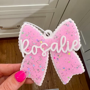 Può includere: Un ornamento a forma di fiocco rosa con un bordo bianco smerlato e dettagli glitterati argentati. Il nome Rosalie è scritto in corsivo bianco in rilievo al centro. Un cordoncino argentato è fissato sulla parte superiore per appenderlo.