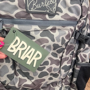 Puede incluir: Una mochila con estampado de camuflaje en tonos grises, marrones y blancos. La mochila tiene cierre de cremallera, un bolsillo lateral y una etiqueta que dice "BRIAR" en letras blancas. Se ve un parche con la palabra "Burlebo" y un gr&aacute;fico de ciervo y pato.