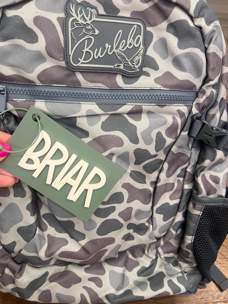 Puede incluir: Una mochila con estampado de camuflaje en tonos grises, marrones y blancos. La mochila tiene cierre de cremallera, un bolsillo lateral y una etiqueta que dice "BRIAR" en letras blancas. Se ve un parche con la palabra "Burlebo" y un gr&aacute;fico de ciervo y pato.