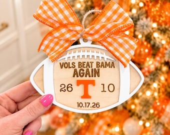 May include: A football-shaped wooden ornament featuring a white border and a natural wood center. It displays the text VOLS BEAT BAMA AGAIN, the numbers 26 and 10, a large orange letter T, and the date 10.17.26. An orange and white gingham bow is attached.