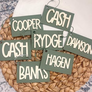Puede incluir: Un conjunto de etiquetas rectangulares verde salvia con nombres en letras blancas en relieve. Cada etiqueta tiene un lazo plateado para colgar. Las etiquetas est&aacute;n dispuestas sobre una superficie tejida de color marr&oacute;n claro.