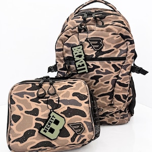 May include: A backpack and a matching lunch bag featuring a tan, brown, and black camouflage pattern. Both items include black zippers, black mesh side pockets, and a black rubberized logo patch reading BECKETT with a stylized letter B.