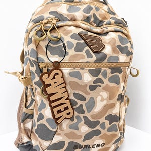 May include: A tan, brown, and grey camouflage backpack featuring a top handle, zippered compartments, and a brown rubberized logo patch. A brown luggage tag with the name SAWYER in raised lettering is attached to the front zipper pull.