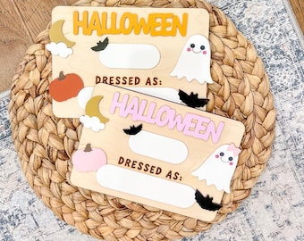 May include: Two wooden Halloween-themed signs. The signs have the word "HALLOWEEN" in orange and pink, with space to write "DRESSED AS:". Each sign has a ghost, bat, pumpkin, moon, and cloud cutouts.