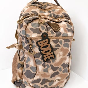 May include: A tan, brown, and grey camouflage print backpack with a top handle, multiple zippered compartments, and side mesh pockets. A black luggage tag with the name BODIE in tan letters hangs from the front zipper. The brand name BURLEBO is printed at the bottom.
