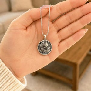 May include: A silver-toned necklace featuring a circular pendant with an embossed profile of a figure wearing a feathered headdress. The word FREYA is inscribed at the bottom of the coin-shaped charm, which hangs from a delicate silver-toned chain.