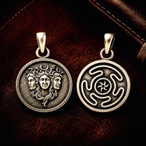 May include: Two circular silver-toned metal pendants with a weathered finish. One pendant features a relief of a three-faced figure with a crescent moon above, while the other displays a central star surrounded by a continuous, winding geometric pattern.