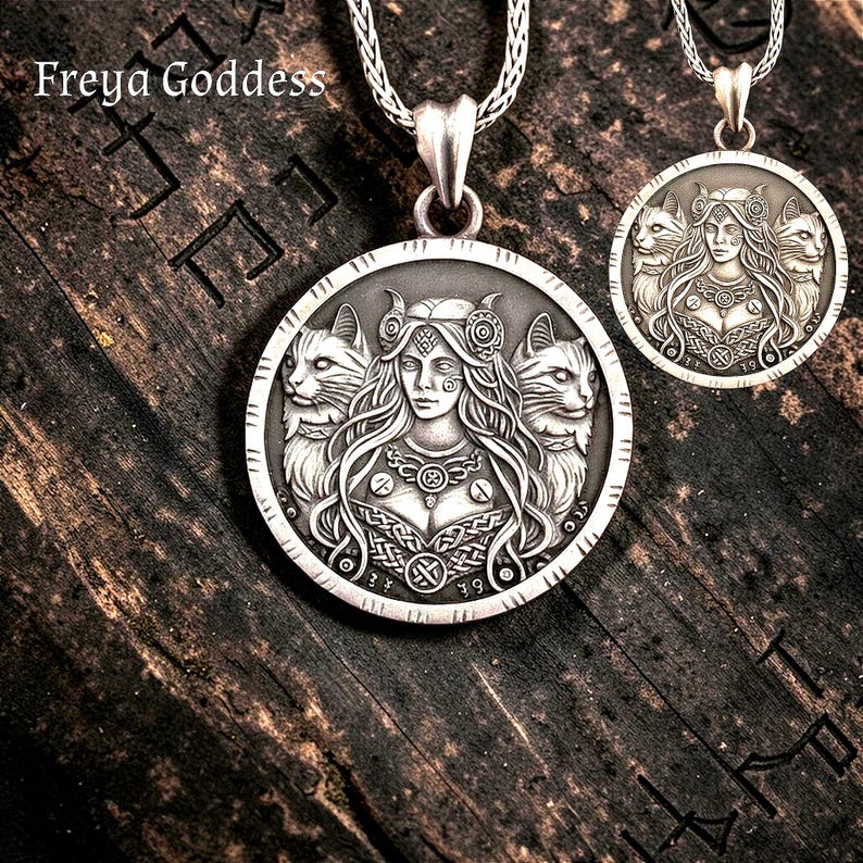 May include: A silver-toned circular pendant necklace featuring an embossed relief of a central figure with long hair flanked by two cats. The pendant is suspended from a silver chain and includes the text Freya Goddess in the upper left corner.