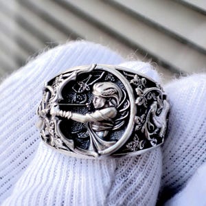 Può includere: Anello d'argento con un rilievo dettagliato di una donna con arco e freccia, circondata da motivi floreali e animali. L'anello ha una finitura antica, che mostra un'intricata lavorazione artigianale e un design classico. L'anello è tenuto da un guanto bianco.
