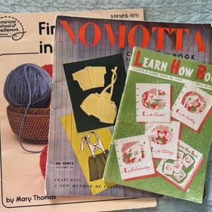 May include: Three vintage instructional craft booklets stacked together. The top booklet is green with the text Learn How Book. The middle booklet is titled Nomotta and features yellow knitwear illustrations. The bottom booklet is beige by Mary Thomas.