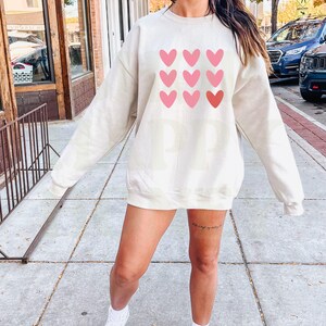 Pode incluir: Uma camisa de moletom de cor creme com um gr&aacute;fico de cora&ccedil;&otilde;es rosa e vermelhos na frente. A camisa de moletom &eacute; de tamanho grande e tem um caimento relaxado.