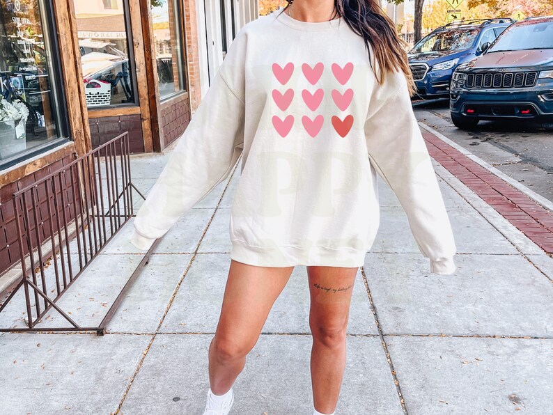 Pode incluir: Uma camisa de moletom de cor creme com um gr&aacute;fico de cora&ccedil;&otilde;es rosa e vermelhos na frente. A camisa de moletom &eacute; de tamanho grande e tem um caimento relaxado.