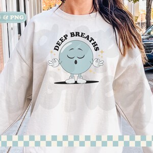 May include: A white sweatshirt with the text "DEEP BREATHS" in black letters. A cartoon blue circle with a face and arms is in the center of the text. The circle has its eyes closed and is holding its hands in a yoga pose.