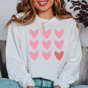 Pode incluir: Camisola branca com um design de cora&ccedil;&otilde;es rosa e vermelho. Os cora&ccedil;&otilde;es est&atilde;o dispostos em um padr&atilde;o de grade.