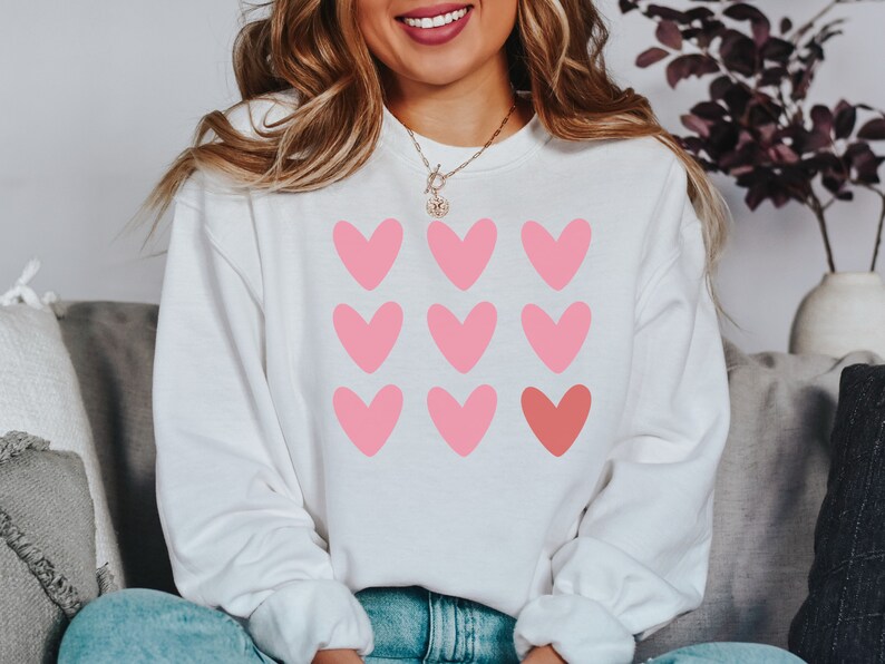 Pode incluir: Camisola branca com um design de cora&ccedil;&otilde;es rosa e vermelho. Os cora&ccedil;&otilde;es est&atilde;o dispostos em um padr&atilde;o de grade.