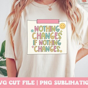 Puede incluir: Una camiseta blanca con un gr&aacute;fico rosa, amarillo, azul y verde que dice "Nada cambia si nada cambia". El gr&aacute;fico est&aacute; en una caja con una cinta rosa en la parte superior y una carita sonriente en la esquina.