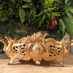 Puede incluir: Centro de mesa decorativo dorado y ornamentado con intrincados adornos y un diseño central de rostro. El recipiente rectangular tiene asas curvas y descansa sobre cuatro patas. El fondo presenta vegetación.