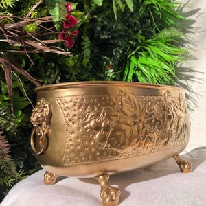 May include: A gold-coloured planter with a detailed relief design. The planter features a lion's head with a ring handle on one side and is supported by three paw-shaped feet. The relief depicts grapes and leaves.