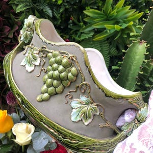 Op de afbeelding: Een decoratieve keramische plantenbak met een grijze basis en groene rand. De plantenbak heeft een reliëfontwerp van groene druiven en bladeren met gouden accenten. De plantenbak is omgeven door groen en bloemen.