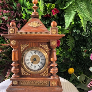 May include: Antique wooden clock with a round face and Roman numerals. The clock has ornate gold-colored accents and a decorative top. The clock is brown with a gold-colored clock face and accents. The clock is a vintage item.