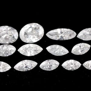 Könnte beinhalten: Eine Auswahl weißer Diamanten in verschiedenen Formen, darunter ovale und marquiseförmige Schliffe, vor einem schwarzen Hintergrund angeordnet. Die Diamanten funkeln und zeigen ihre Facetten und Brillanz. Die Kollektion umfasst mehrere ovale und marquiseförmige Steine.