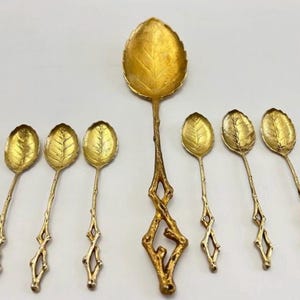 Puede incluir: Un juego de seis cucharas doradas con cuencos en forma de hoja y mangos en forma de ramita. Una cuchara más grande presenta un diseño detallado de hoja y un mango decorativo. Las cucharas están dispuestas sobre una superficie blanca.