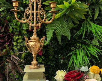 Puede incluir: Un candelabro dorado con tres candeleros, detalles ornamentados y una base decorativa. El candelabro se encuentra sobre una base de piedra verde y blanca con patas doradas. El fondo presenta una variedad de plantas y flores verdes.