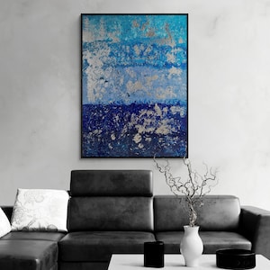 Puede incluir: Pintura abstracta con tonos azules y plateados. La pintura está enmarcada en negro y cuelga de una pared blanca sobre un sofá de cuero negro.