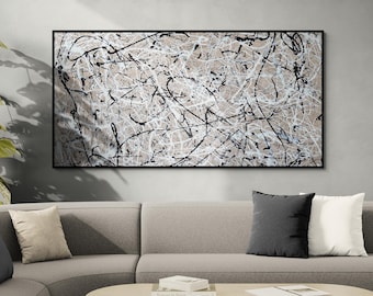 May include: Abstract art piece in a black frame. The artwork features a beige background with black and white paint splatters and lines. The painting hangs above a gray sofa with pillows. A plant and a wooden door are also visible.