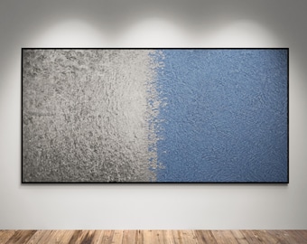 May include: Abstract wall art featuring a textured surface divided into two halves. One side is a light gray, the other is a medium blue. The artwork is framed in black and hangs on a white wall above a wooden floor.