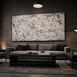 Puede incluir: Una pintura abstracta con salpicaduras de pintura negra y blanca cuelga sobre un sofá gris en una sala de estar moderna. La habitación cuenta con una pared de ladrillo, una mesa de centro de madera oscura y sillones de cuero. La iluminación es tenue.
