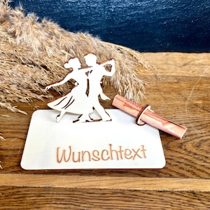 Könnte beinhalten: Ein rechteckiger Sockel aus hellem Holz mit einer lasergeschnittenen Silhouette eines tanzenden Paares und dem eingravierten Text "Wunschtext". Ein kleiner Holzring hält ein zusammengerolltes Papier mit sichtbarem Text neben den Figuren.