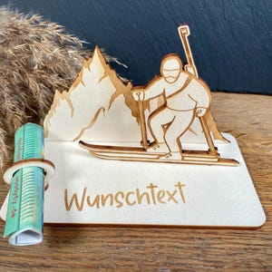 Könnte beinhalten: Ein heller Holzaufsteller mit einer lasergeschnittenen Silhouette eines Skifahrers vor Berggipfeln. Der Sockel enthält eine kreisförmige Halterung für gerolltes Papier und den eingravierten Text "Wunschtext" auf der Vorderseite.