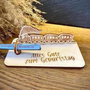 Könnte beinhalten: Ein heller Holzaufsteller mit einer lasergeschnittenen Zug-Silhouette inklusive Rauch. Auf der Basis ist der Text Alles Gute zum Geburtstag eingraviert. Ein blaues rechteckiges Teil mit der Aufschrift Spielgeld woodyfy.de ist befestigt.