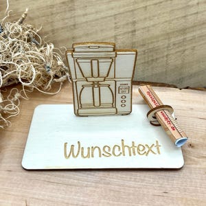 Könnte beinhalten: Ein heller Holzausschnitt einer Kaffeemaschine auf einem rechteckigen Sockel mit dem eingravierten Text "Wunschtext". Ein kleiner Holzzylinder mit dem aufgedruckten Text "spielgeld woodyty.de" ist an der Seite des Sockels befestigt.