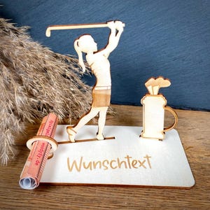 Könnte beinhalten: Ein heller Holzaufsteller mit einer ausgeschnittenen Golferfigur in Schwungposition und einer Golftasche. Der Sockel enthält eine kreisförmige Halterung für einen gerollten Gegenstand sowie den eingravierten Text "Wunschtext" in Schreibschrift.