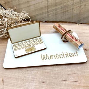 Könnte beinhalten: Eine weiße rechteckige Holzplatte mit dem in Gold eingravierten Wort Wunschtext, darauf ein kleines Laptop-Modell aus Holz und eine gerollte Papierrolle, die von einem Holzring gehalten wird. Die Rolle trägt den roten Text Spielgeld und woodyfy.de.