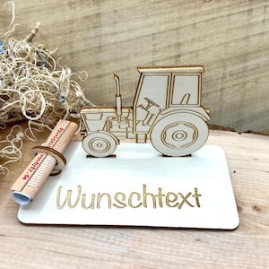Könnte beinhalten: Ein heller Holztraktor-Ausschnitt auf einem rechteckigen Sockel mit dem lasergravierten Text "Wunschtext". Ein kleiner Holzzylinder mit dem aufgedruckten Text "woodyfy.de" ist an der Seite des Sockels befestigt und dient als Halterung.