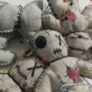 Puede incluir: Un grupo de cinco muñecos vudú hechos de tela blanca con costuras negras y botones negros. Los muñecos están decorados con corazones rojos, X negras y tienen alfileres clavados en ellos.