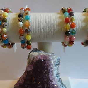 Könnte beinhalten: Vier bunte Perlenarmbänder mit verschiedenen Steinen und Metallakzenten, die auf einem weißen Ständer ausgestellt sind. Die Armbänder sind aus verschiedenen farbigen Steinen gefertigt, darunter rot, orange, gelb, grün, blau und lila. Der Ständer besteht aus einem weichen, weißen Stoff.