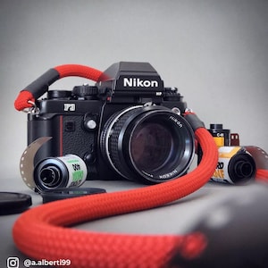 Könnte beinhalten: Eine schwarze Nikon F3 Kamera mit rotem Gurt und Objektiv. Das Nikon-Logo ist oben und auf dem Objektiv zu sehen. Filmrollen und ein Objektivdeckel sind ebenfalls vorhanden. Das Bild hat eine Vintage-Ästhetik.