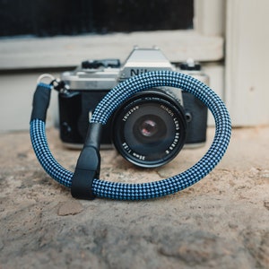 Può includere: Un cinturino per fotocamera blu e nero con un motivo a scacchi bianchi. Il cinturino è avvolto attorno a una fotocamera argentata con un obiettivo nero.