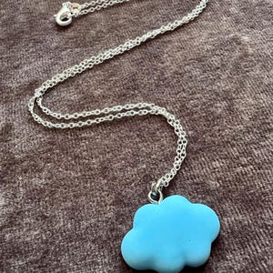 Puede incluir: Un collar de cadena de metal plateado con un colgante liso en forma de nube de color azul claro. El collar incluye un cierre de mosquetón estándar en el extremo de la cadena, lo que proporciona una forma segura de abrochar la pieza alrededor del cuello.