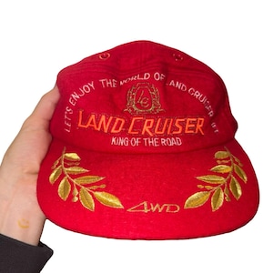 Op de afbeelding: Een rode honkbalpet met geborduurde tekst "LET'S ENJOY THE WORLD OF LAND CRUISER" en "KING OF THE ROAD" in het wit, met "LAND CRUISER" in het oranje. Het bevat een goudkleurig logo, gouden laurierbladborduursels en de tekst "4WD".