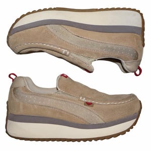 May include: Two tan suede slip-on shoes with a platform sole. The shoes have a beige and gray sole, with a red accent on the heel and tongue. The shoes have a stitched design and a small red heart logo.