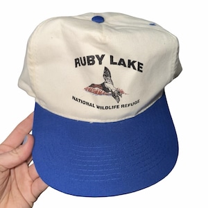 May include: A white and blue baseball cap featuring the text "RUBY LAKE NATIONAL WILDLIFE REFUGE" and a bird graphic. The cap has a blue peak and a blue button on the crown.